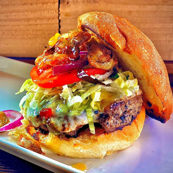 A towering Main Event Grill burger with melted cheese, lettuce, tomato, onions, and sauce.